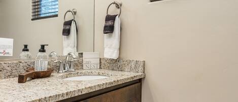 A bathroom with a granite countertop, dark wooden cabinets, a large mirror, and a window with blinds.