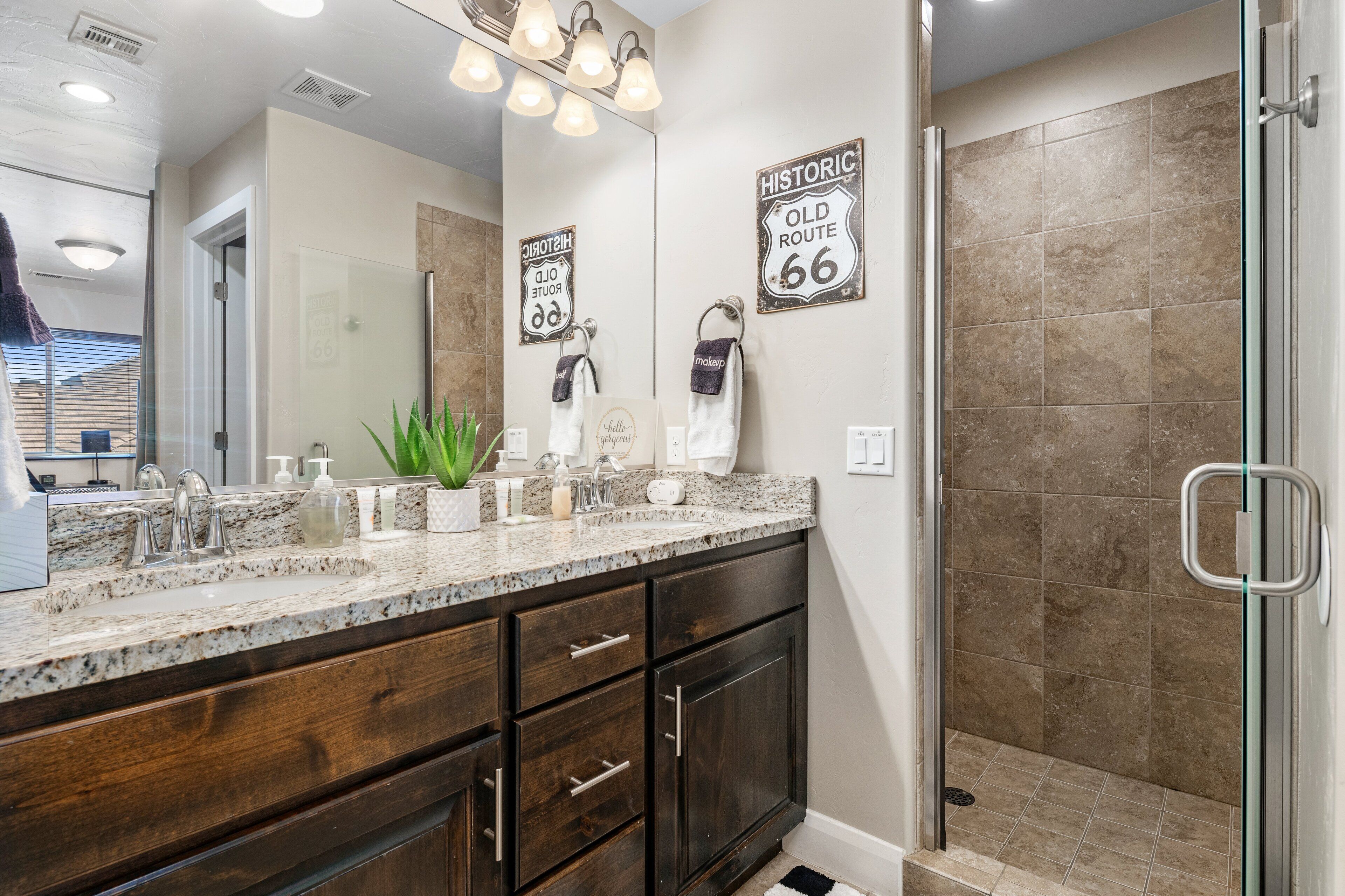 A bathroom with a granite countertop, dark wood cabinets, a large mirror, and a shower with beige tiles.