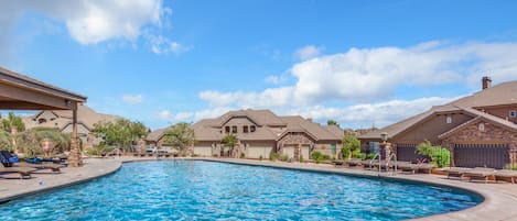Piscina con sillones de descanso y una zona techada, rodeada de casas bajo un cielo despejado.