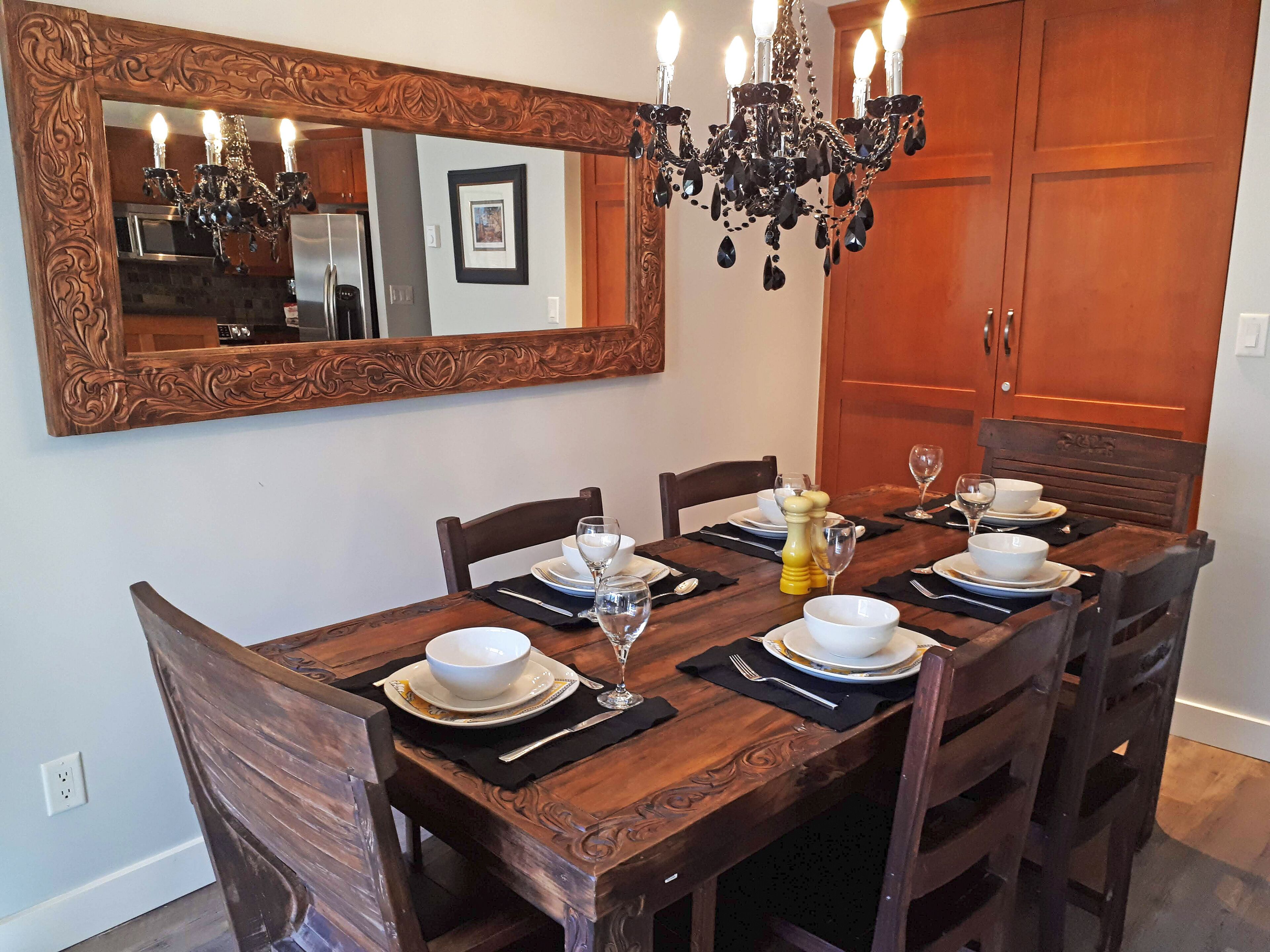 A dining room with a wooden table set for four, a chandelier, and a large mirror above the table.