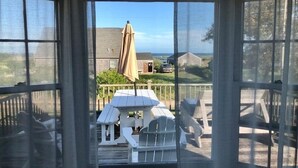 A view through a screened door to an outdoor deck with a table and chairs, a covered umbrella, and a view of a residential area with houses and a car.