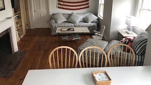 A cozy living room with a striped sofa, a wooden coffee table, and a dining table with chairs. An American flag hangs on the wall, and a fireplace is visible in the corner.
