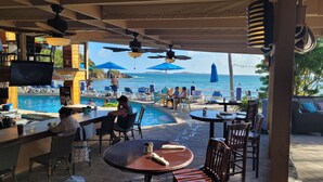 An outdoor pool area with lounge chairs and umbrellas, a bar with a television, and a view of the sea. - REMODELED 2026! Prime OCEANFRONT POOLSIDE 2BR/2BA Condo-BEST location+Generator (St Thomas)