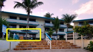 A multi-story building with a blue roof, a staircase leading to the entrance, and palm trees. - REMODELED 2026! Prime OCEANFRONT POOLSIDE 2BR/2BA Condo-BEST location+Generator (St Thomas)