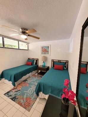 A hotel room with two beds, a ceiling fan, a window with blinds, a painting, a nightstand, and a floral rug. - REMODELED 2026! Prime OCEANFRONT POOLSIDE 2BR/2BA Condo-BEST location+Generator (St Thomas)
