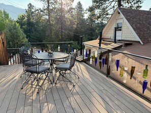 A wooden deck with a round table and chairs, a small house with a sloped roof, and a view of trees and mountains in the background.