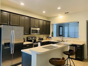 A modern kitchen with dark cabinets, stainless steel appliances, a central island with stools, and a window with blinds.