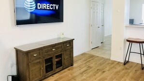 Un salon moderne avec un téléviseur à écran plat fixé au mur, diffusant les chaînes de DIRECTV, un meuble en bois avec des tiroirs, un petit tabouret et une porte donnant sur une autre pièce.