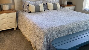 Une chambre à coucher comprenant un lit, deux tables de chevet, une commode, une œuvre d’art représentant une carte, un ventilateur de plafond et une fenêtre avec des stores.