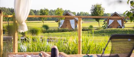 Una terraza de madera con una pareja relajándose en un banco, con vistas a un campo cubierto de césped y dos cabañas de madera.