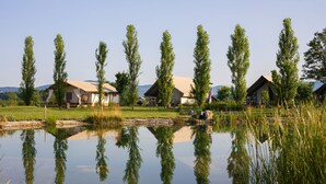 A tranquil lakeside setting with several cabins, a clear reflection in the water, and a grassy area with trees. - Glamping Kolpa Resort (Metlika)