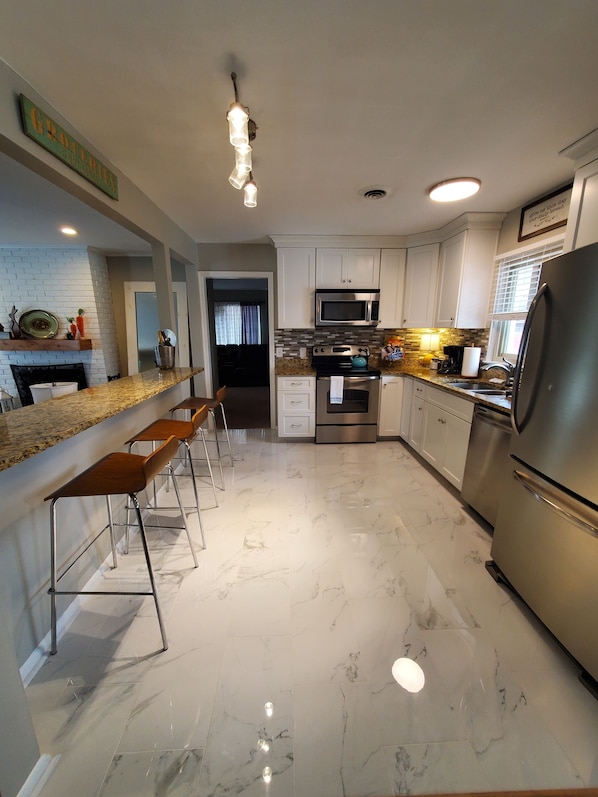 Una cocina moderna con armarios blancos, un refrigerador de acero inoxidable, un microondas y una estufa. Hay taburetes altos a lo largo de la encimera y una pared de ladrillo con un estante arriba.