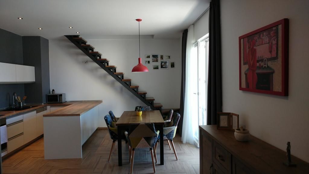 A modern kitchen with a wooden island, a dining area with a table and chairs, a staircase, and a red pendant light.