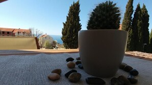 Blick von einem Balkon mit einer großen weißen Schale auf einer Matte, umgeben von Steinen, mit Ausblick auf eine Küstenlandschaft mit Gebäuden und hohen Bäumen.