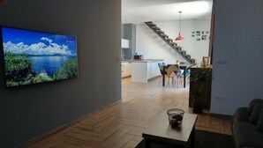 Ein modernes Wohnzimmer mit Holzboden, einem an der Wand befestigten Fernseher, einem Essbereich mit Stühlen und einer Treppe, die zu einer weiteren Ebene führt.