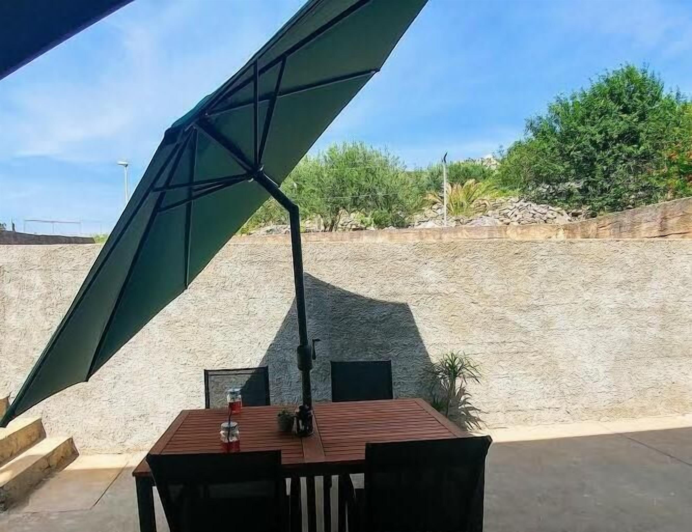 Un patio con mesa y sillas bajo un gran paraguas, rodeado por un muro de piedra y follaje.