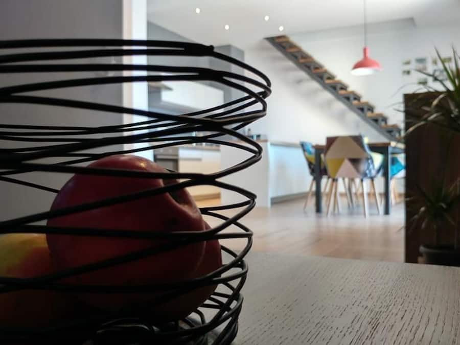 Una cocina moderna con una mesa de madera, una cesta metálica negra con manzanas y una escalera que sube a un nivel superior.