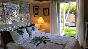 A bedroom with a stone wall, a bed with palm tree pillows, a nightstand with a lamp, and a sliding glass door to a garden. - Hale Alaula - Ocean View: Private Home Near Kona Town w/Spa, AC, Spacious Lanai (Kailua Kona)
