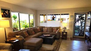 A living room with a sectional sofa, a coffee table, and a rug. There are two lamps, a door with a decorative glass panel, and a window with blinds. - Hale Alaula - Ocean View: Private Home Near Kona Town w/Spa, AC, Spacious Lanai (Kailua Kona)