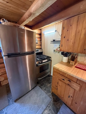 A compact kitchen with a stainless steel refrigerator, a gas stove, wooden cabinets, and a stone-tiled floor.