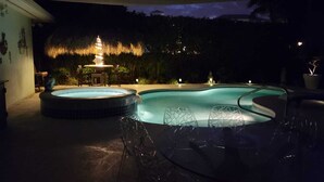 Una piscina con hidromasaje y sillas de descanso, rodeada por una terraza con techo de paja e iluminación ambiental.