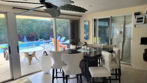 Comedor con una mesa grande y sillas blancas, ventilador de techo y vista a una piscina y asientos exteriores.