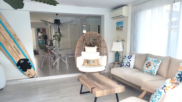 Una sala de estar moderna con un sofá blanco, una mesa de centro de madera, una silla de mimbre y una tabla de surf apoyada en la pared.