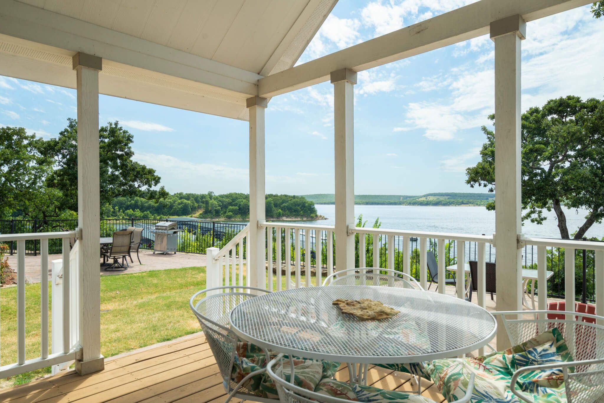 A covered outdoor area with a view of a lake, featuring a round metal table with chairs and a grill.