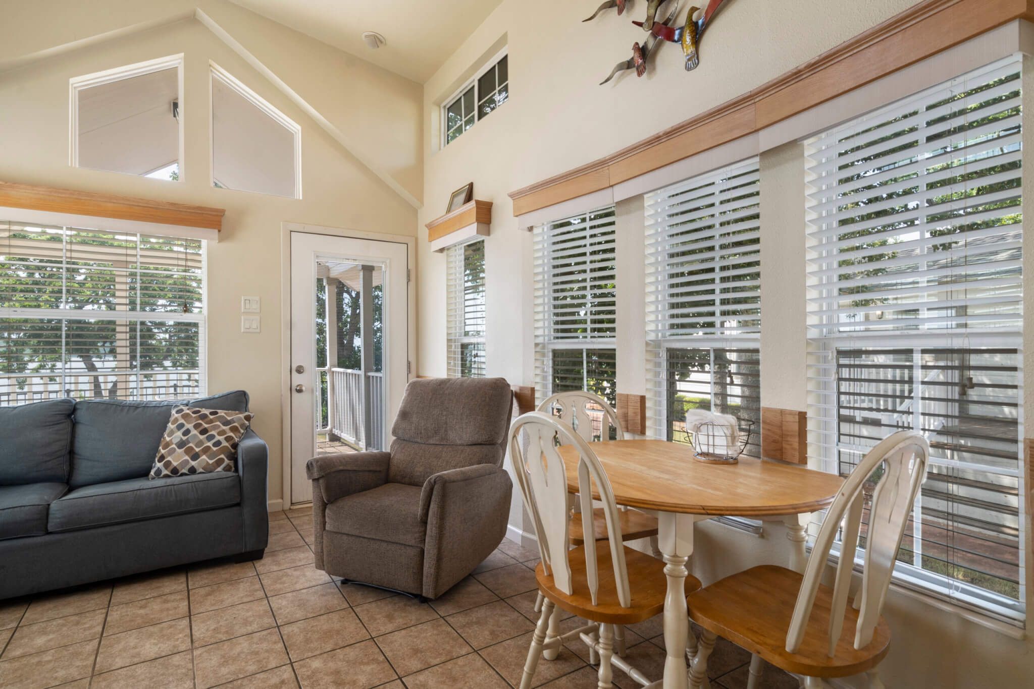 A spacious living room with a gray sofa, a wooden dining table with white chairs, and large windows with blinds.