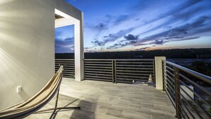 Un balcon moderne avec un hamac, une balustrade et une vue sur un coucher de soleil au-dessus d’une ville.