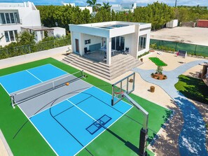 Una cancha de tenis con red, un aro de baloncesto y un pequeño patio con escalones que llevan a una zona de estar cubierta.