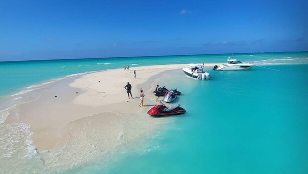 Une plage aux eaux turquoise et limpides, un rivage sablonneux, et quelques personnes et bateaux.