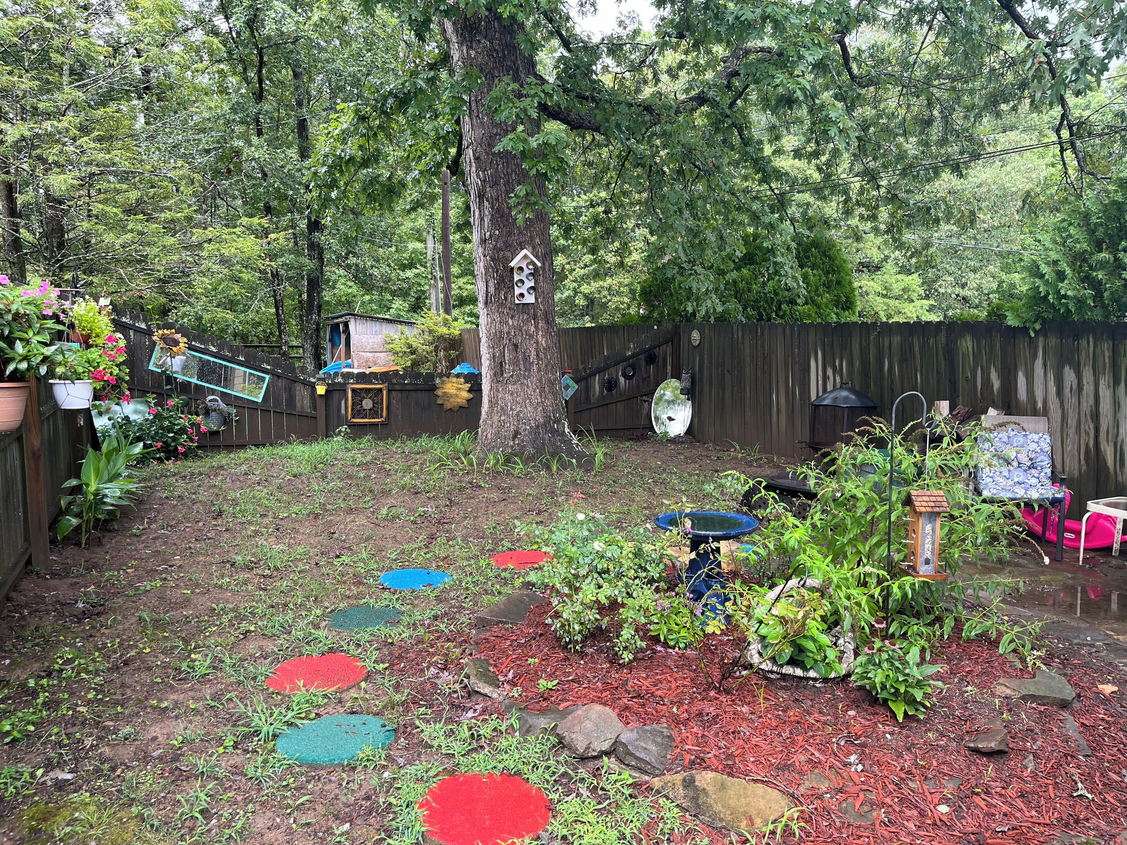 A backyard with a wooden fence, a tree, and various plants and decorations.