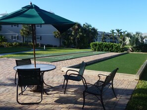A patio area with a table and chairs, a green umbrella, and a shuffleboard court. - Luxury 3 bed  Resort Style Amenities (Naples)