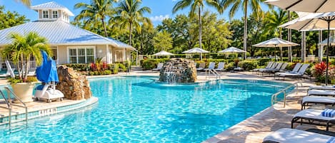 A swimming pool with lounge chairs and umbrellas, surrounded by palm trees and a building with a blue roof.