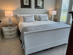 A bedroom with a white bed, two bedside tables, and a nightstand. There are framed pictures on the wall and a window with blinds. - Luxury 3 bed  Resort Style Amenities (Naples)