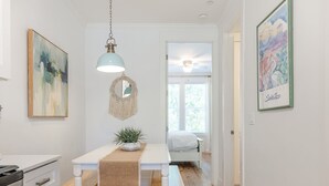Et lyst, hvidt interiør med et tekøkken, et spisebord med en plante og en seng i baggrunden.