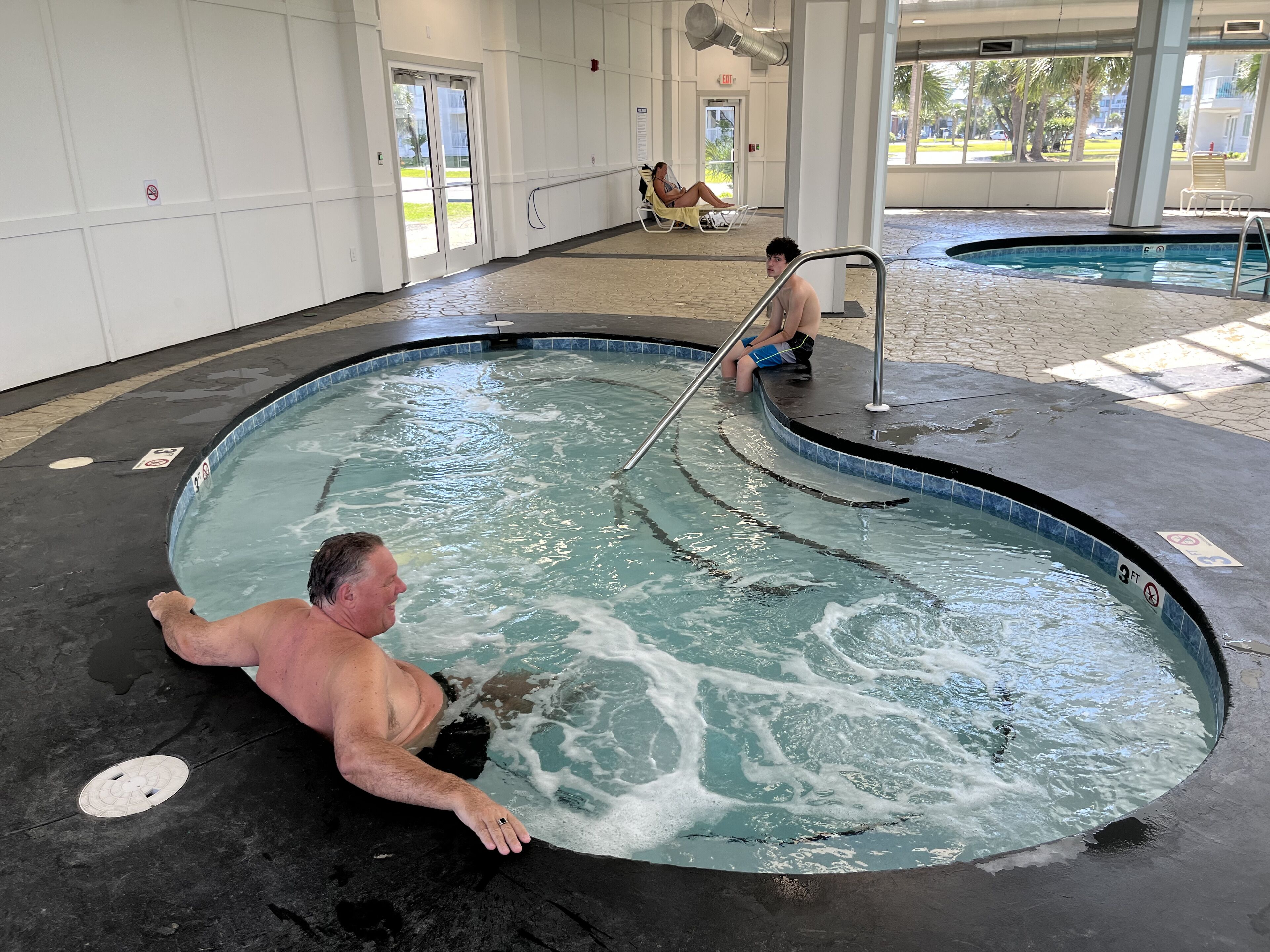 Een man ontspant in een jacuzzi, met een zwembad op de achtergrond.