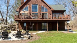 A two-story log cabin with a balcony, stone foundation, and outdoor seating area. - 4BR Lodge Near Lake Geneva - Sitting Pool, Hot Tub, Sauna, Lake Views on 3 Acres (Lake Geneva)
