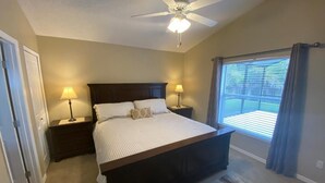 A bedroom with a bed, two bedside lamps, a ceiling fan, a window with curtains, and a view of a deck. - GORGEOUS POOL HOME IN QUIET TRANQUIL SETTING CLOSE TO ATTRACTIONS & OUTLETS (Kissimmee)