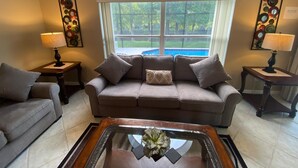 A living room with a beige sofa, two side tables with lamps, a glass coffee table with a decorative item, and a large window with a view of a swimming pool and greenery. - GORGEOUS POOL HOME IN QUIET TRANQUIL SETTING CLOSE TO ATTRACTIONS & OUTLETS (Kissimmee)