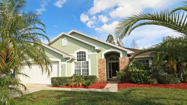 A two-story house with a green facade, a brick entrance, and a well-maintained front yard with red mulch and palm trees. - GORGEOUS POOL HOME IN QUIET TRANQUIL SETTING CLOSE TO ATTRACTIONS & OUTLETS (Kissimmee)