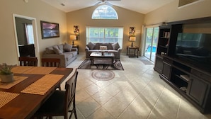 A spacious living room with a high ceiling, a ceiling fan, a large TV, a dining table with chairs, and a sofa. - GORGEOUS POOL HOME IN QUIET TRANQUIL SETTING CLOSE TO ATTRACTIONS & OUTLETS (Kissimmee)