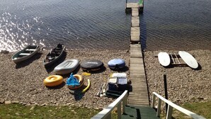 En träbrygga som leder ut till en lugn sjö, med båtar och andra vattenfarkoster längs stranden.