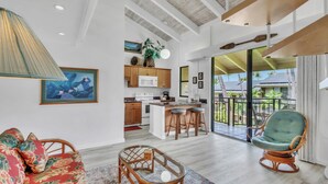 A spacious living area with a sofa, wicker chairs, a glass coffee table, and a kitchen in the background. - 🌴Top floor with ocean  views. Well equipped. AC and WiFi🌴 (Kailua-Kona)