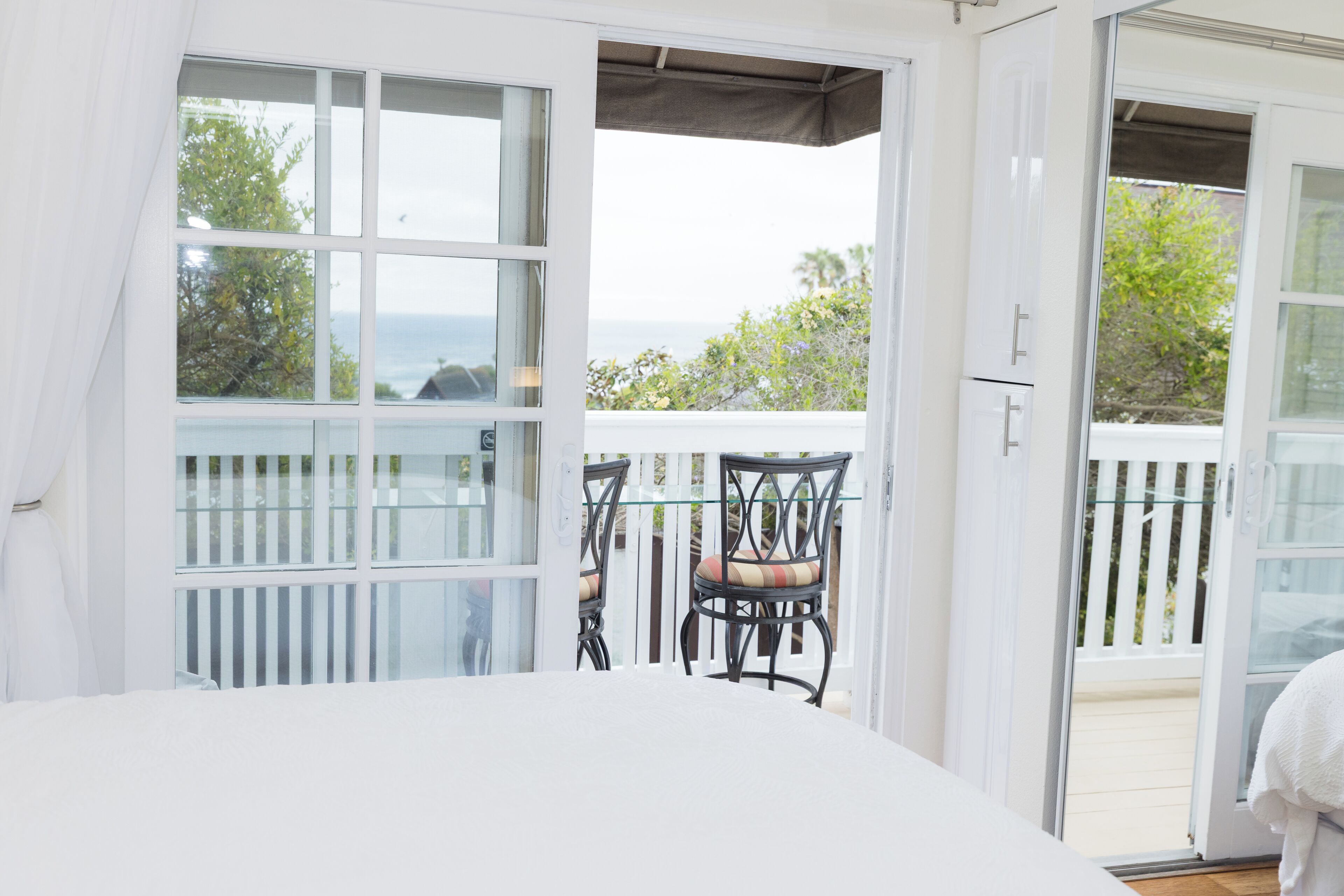 A room with a white bed, a glass door leading to a balcony with a view of the sea, and two chairs with cushions.