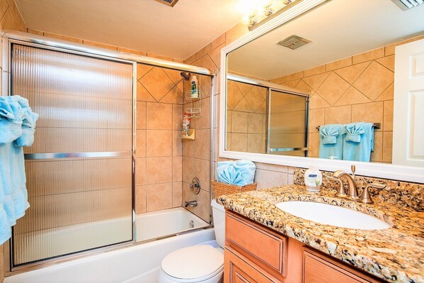 A bathroom with a glass shower enclosure, a white toilet, a sink with a granite countertop, and a large mirror.