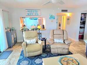 A living room with two beige armchairs, a coffee table, a blue patterned rug, a kitchen area with a blue countertop, and a sign that reads 'SIESTA LIVING THE DREAM KEY FL.' - ON Siesta Key BEACH! Pool,Remodeled! 2bd2ba, sleeps 6, 3mins from door to sand   (Sarasota)