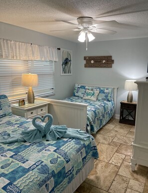 Ein Schlafzimmer mit zwei Betten, einem Deckenventilator, einem Fenster mit Jalousien, einer Wand mit einem maritimen Schild und einem kleinen Tisch mit einer Lampe.