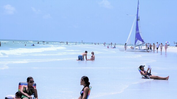 A beach with people sunbathing and a sailboat in the water.
